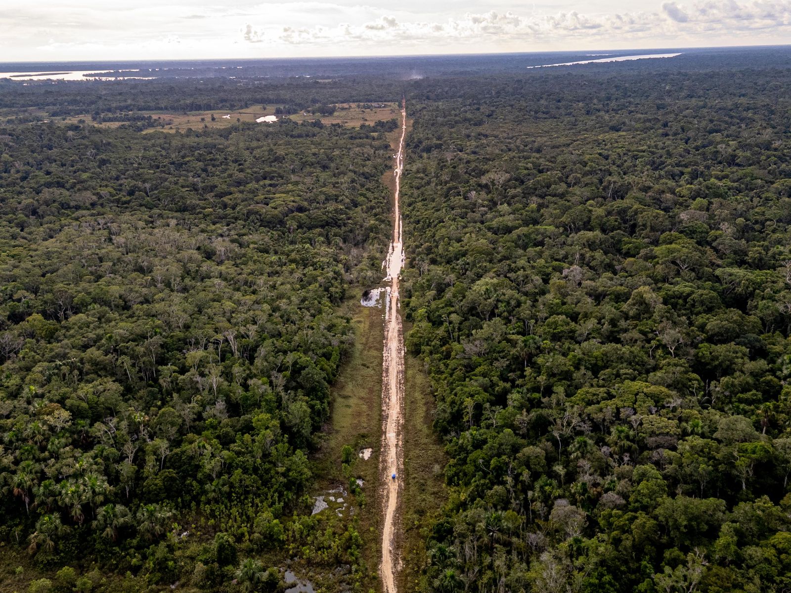 A straight dirt road with vehicles cutting through the forest toward the horizon
